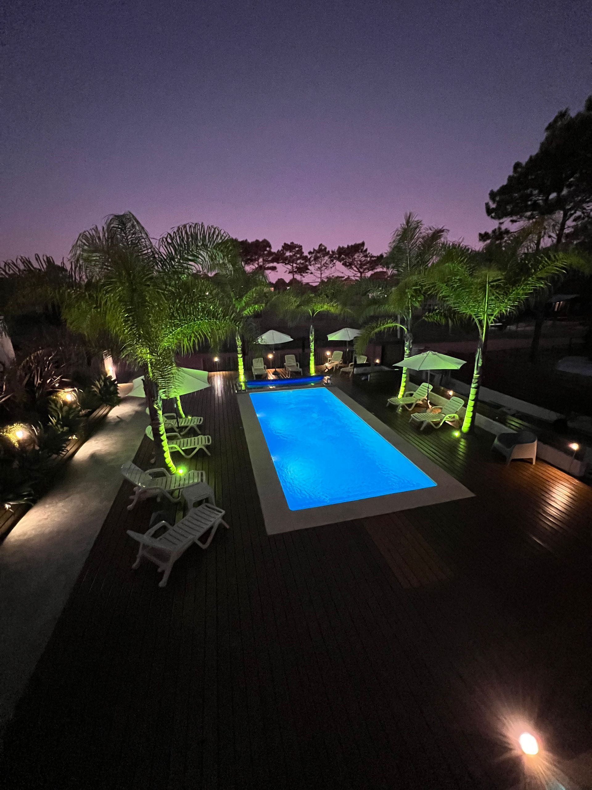 A large swimming pool is lit up at night with umbrellas and chairs around it.