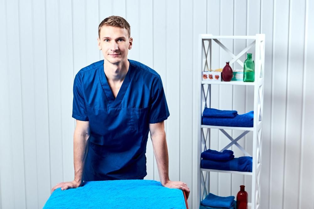 A Man In A Blue Scrub Is Standing Next To A Blue Table — Mark Coulton Cardiff Physiotherapy Centre In Cardiff, NSW