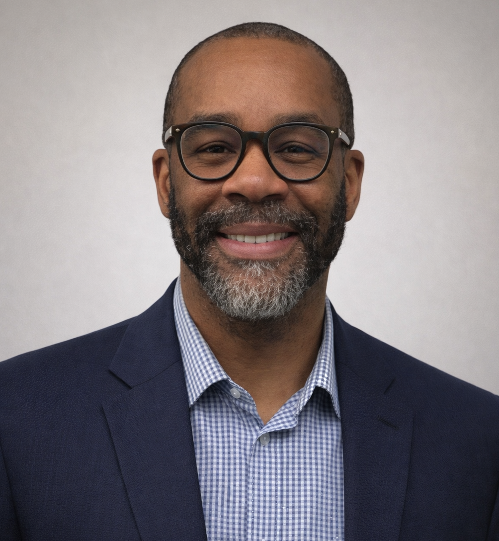 Man wearing glasses, black suit, and blue tie, smiling against a gray background.