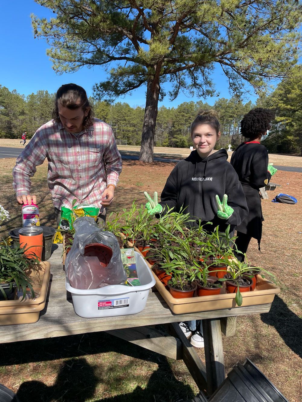 A group of people are standing around a picnic table with potted plants.