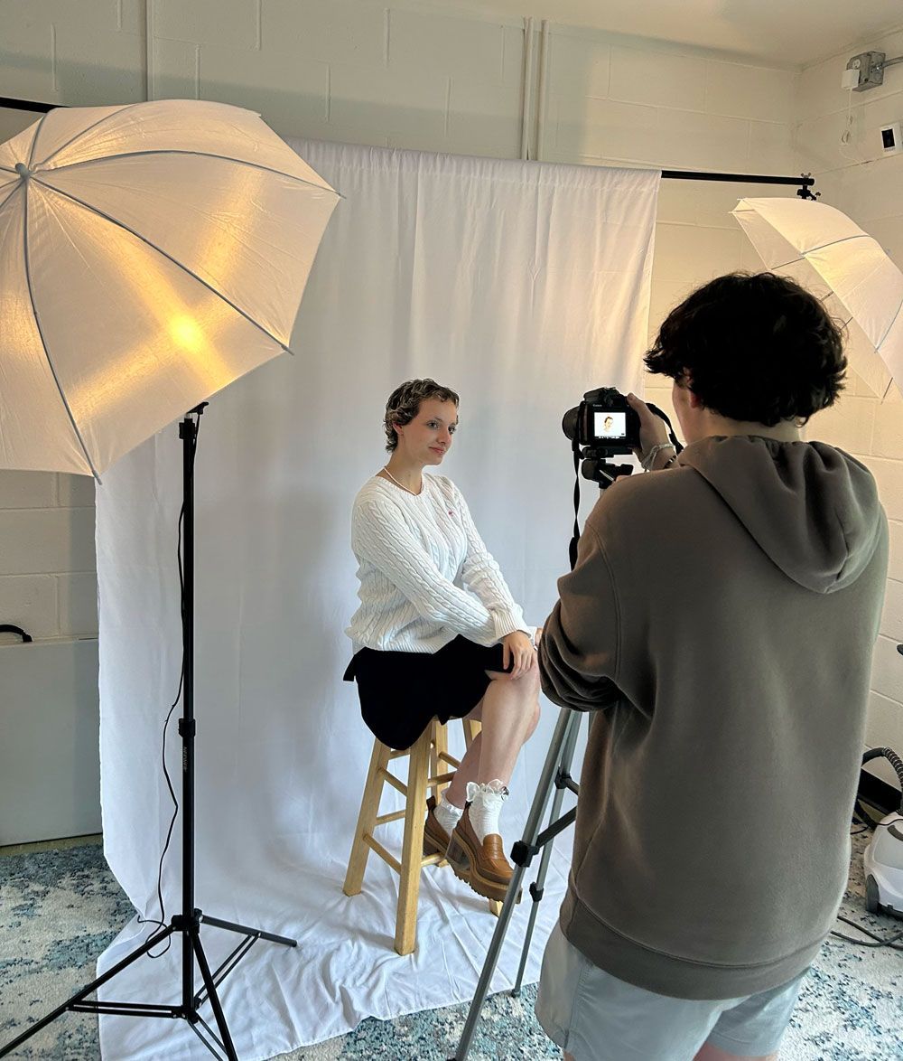 A man is taking a picture of a woman sitting on a stool.
