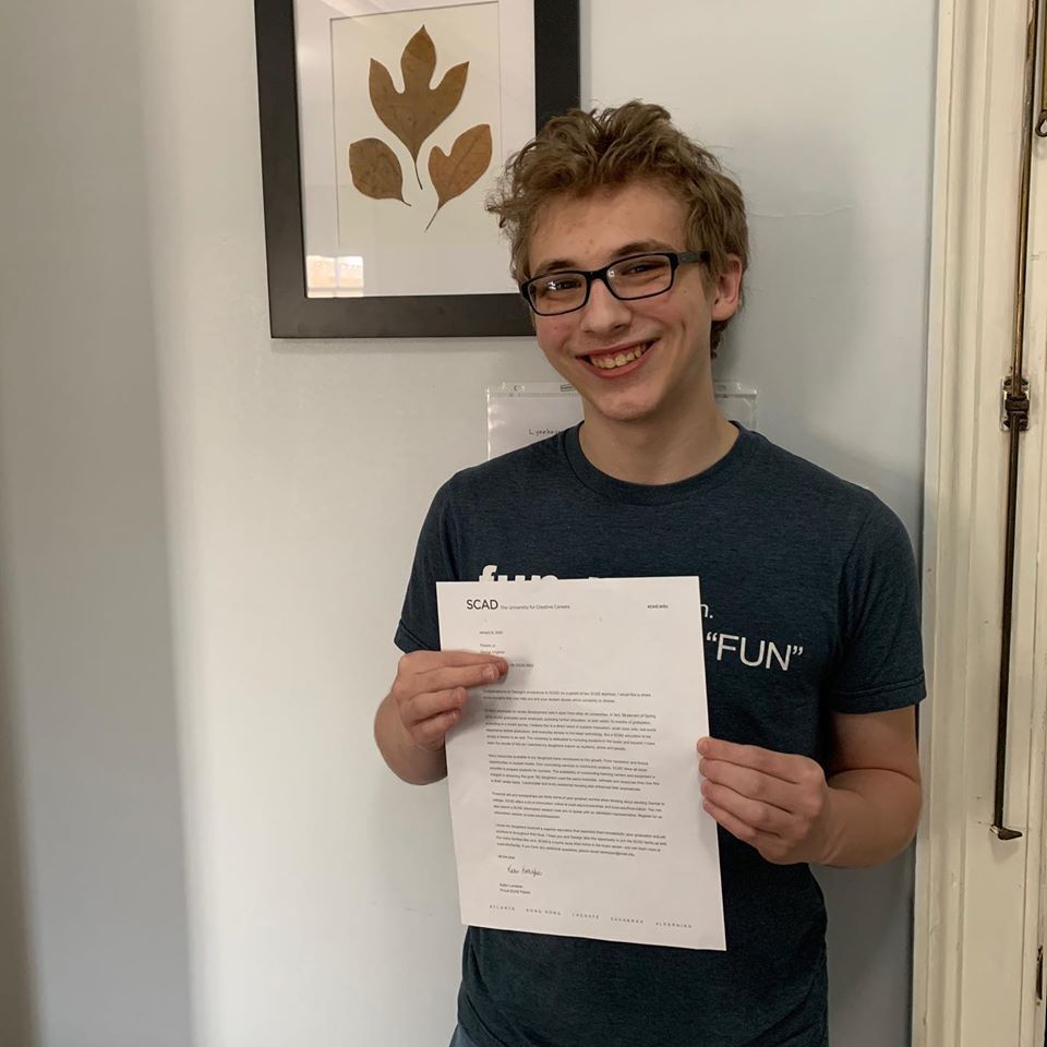 A young man wearing glasses is holding a piece of paper and smiling