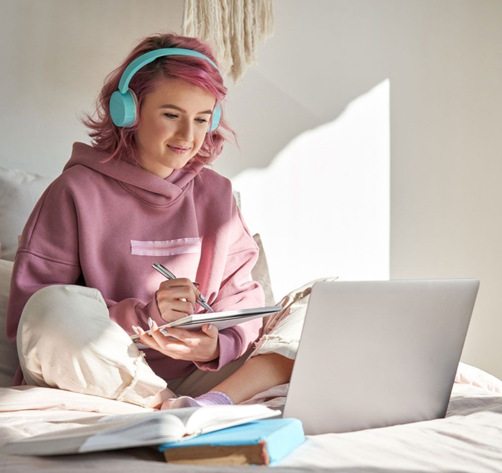 A woman wearing headphones is sitting on a bed using a laptop computer.