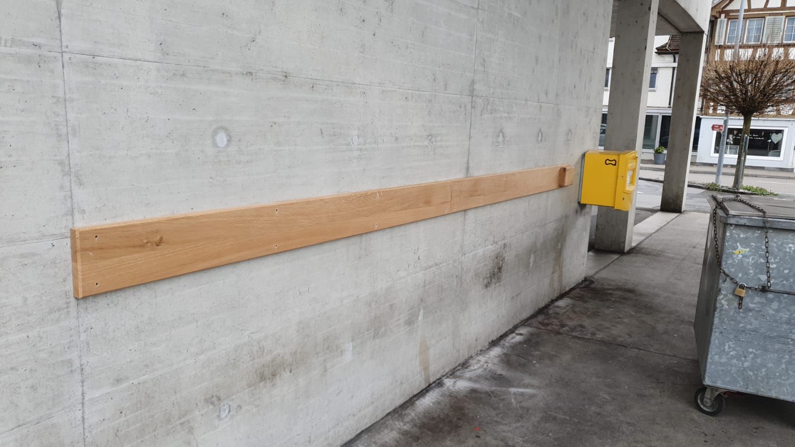 Neben einer Mülltonne ist an einer Betonwand ein Holzgeländer angebracht.