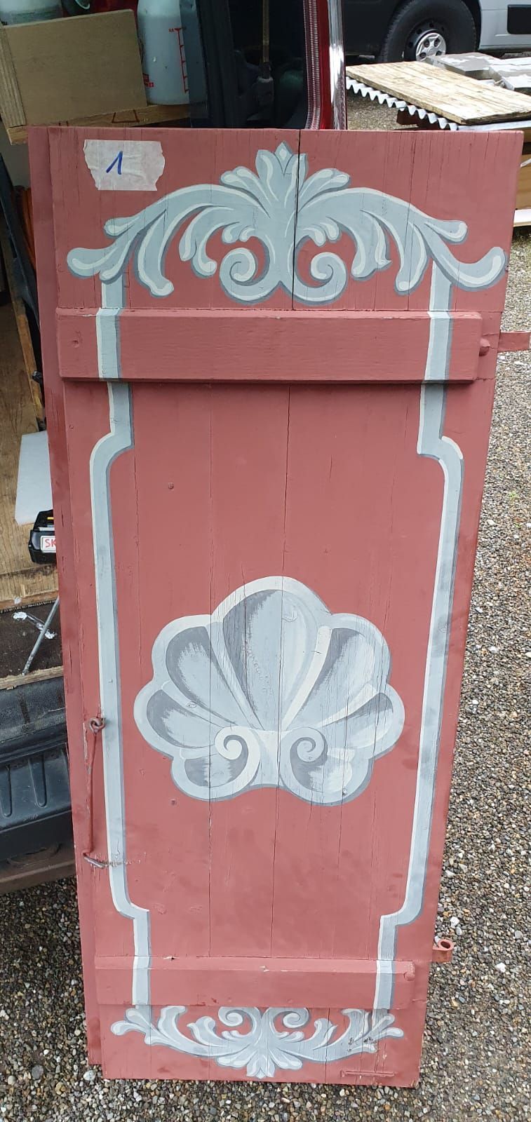 Ein roter Fensterladen mit einem weissen Muster steht auf einer Schotterstrasse.