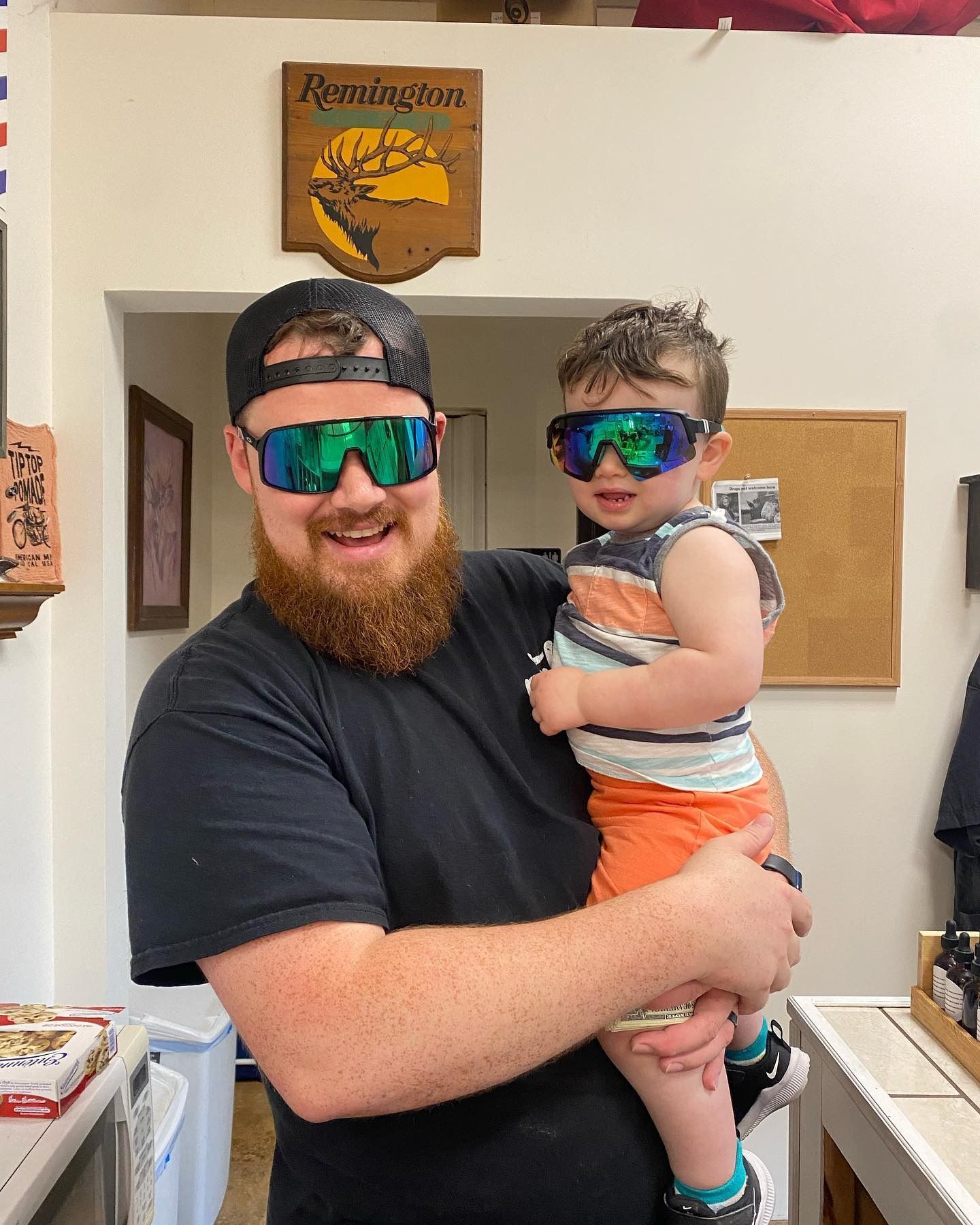 A man with a beard is holding a baby wearing sunglasses.
