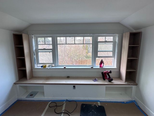 A room with a large window and built-in bench. Bookshelves flank the window. Construction is in progress.