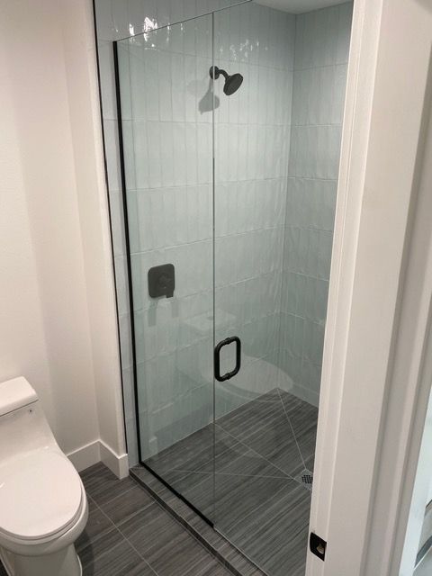 Shower stall with glass door, black hardware, gray tile, and a toilet to the left.