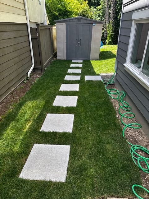 Concrete stepping stones in a grassy yard lead to a gray shed between two buildings. Green hose coiled.