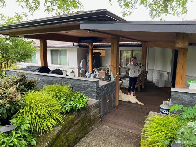 A person and dog on a wooden deck with an attached covered patio. Green plants and a stone wall are in the foreground.