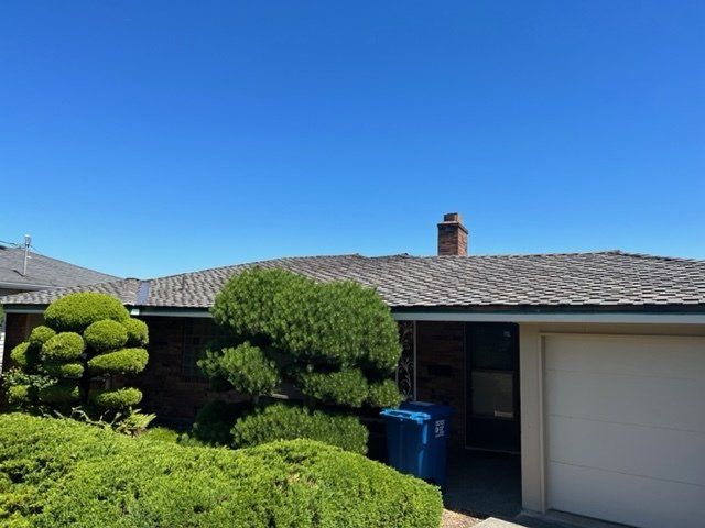 House with a gray roof, brick chimney, and garage door on a sunny day with blue sky. Green shrubs in front.