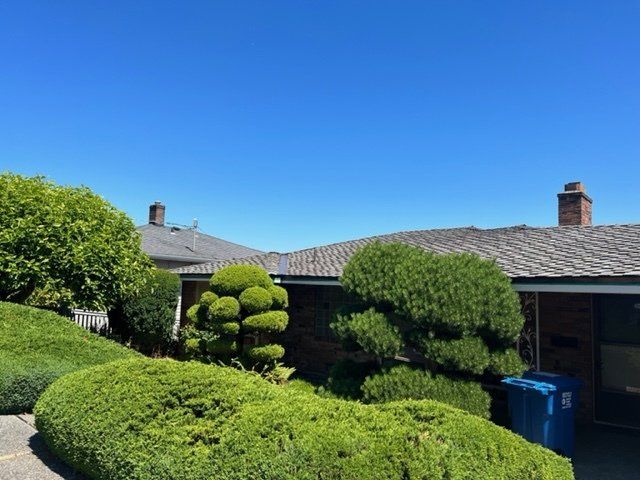 Blue sky over a house with manicured green bushes and a blue trash bin.