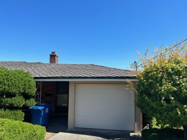 Garage of a brick house with a brown roof and a blue trash bin under a bright blue sky.
