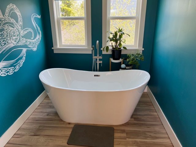 A white freestanding bathtub sits in a teal-walled bathroom with wood-look flooring. Windows and plants are behind the tub.