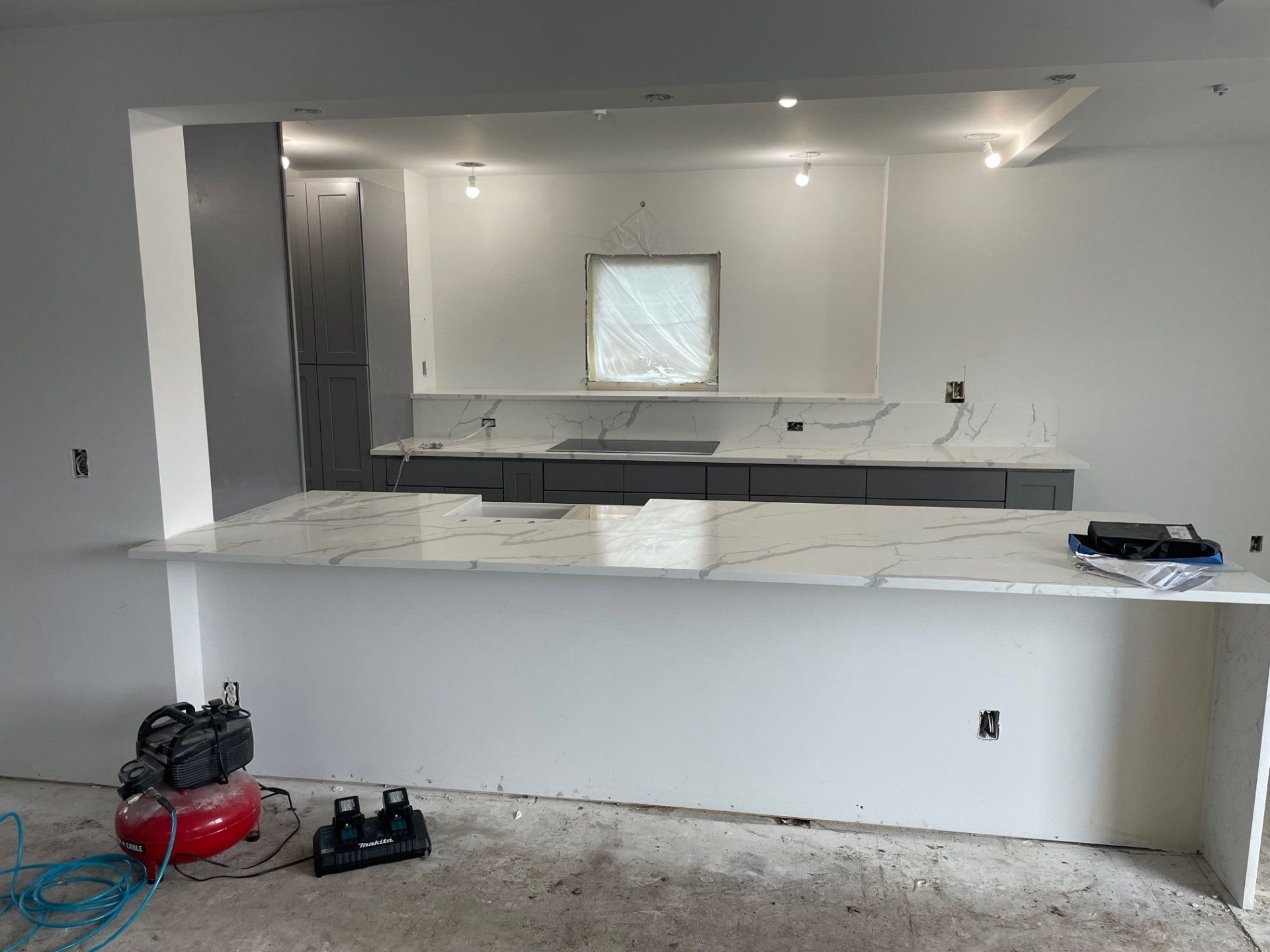 Kitchen under construction with white countertops, gray cabinets, and a pass-through to a second room.