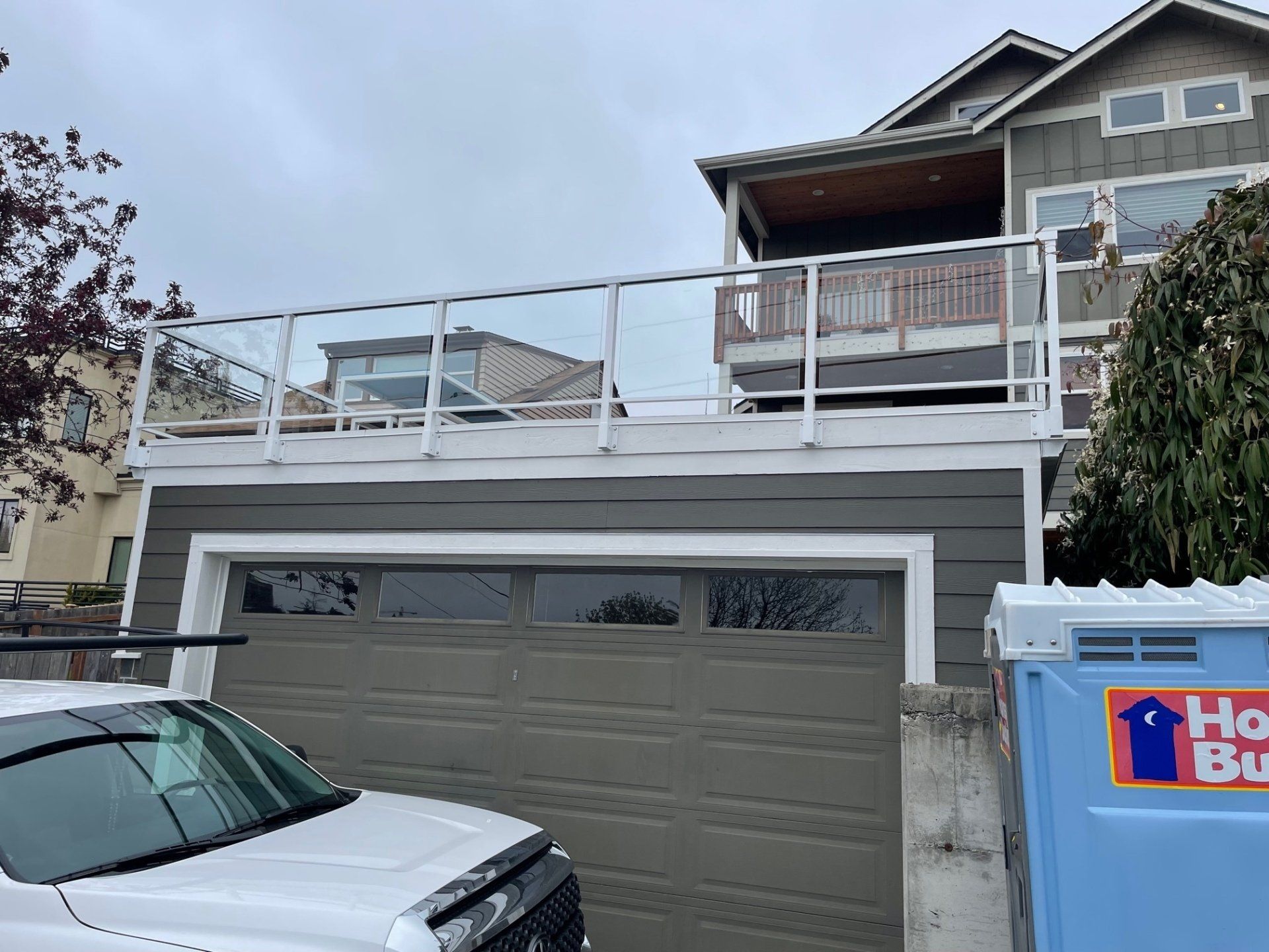 Garage with a glass-railed deck on top, attached to a two-story house with green siding, on a cloudy day.