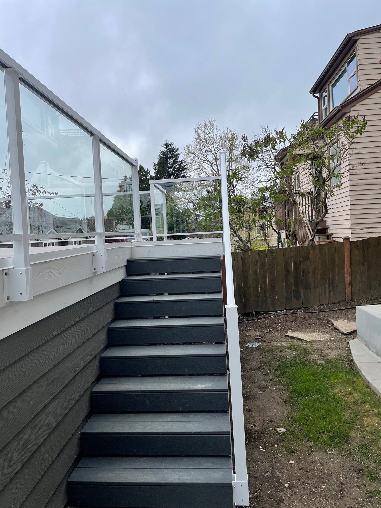 Gray outdoor stairs leading to a deck with glass railing, next to a wood-planked fence.