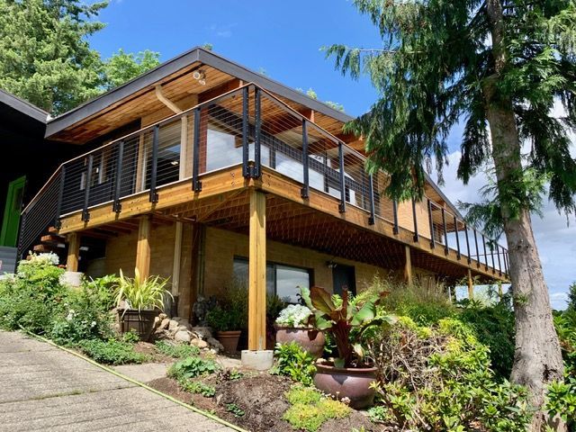 Two-story house with wooden deck, black railings, and lush landscaping. Overcast sky.