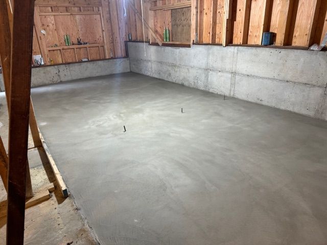 Newly poured concrete floor in a basement. Wood studs and concrete walls surround the space.