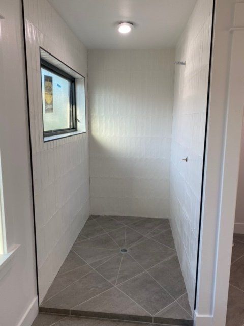 Empty walk-in shower with white tile walls and gray tile floor. Small window and recessed lighting.