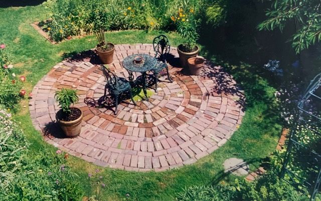 Circular brick patio with black table and chairs, surrounded by grass and potted plants.