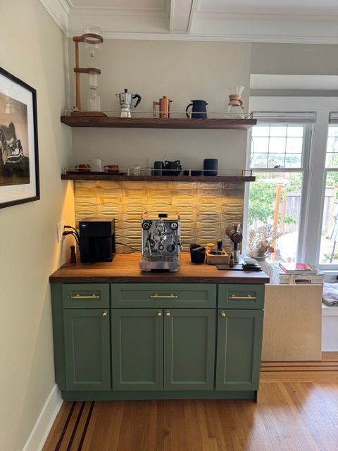 Green coffee bar with espresso machine, wooden shelves with coffee equipment, and green cabinets.