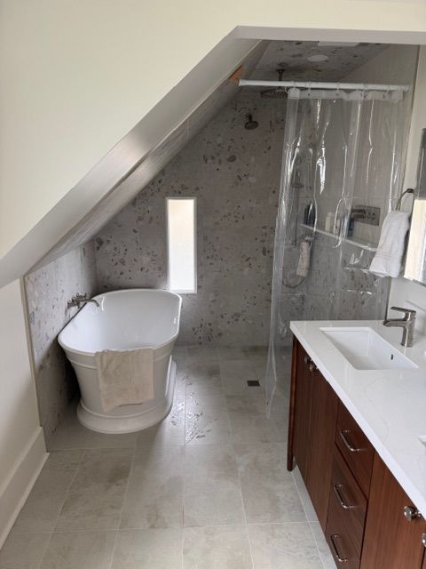 Bathroom with angled ceiling; freestanding tub near small window, shower, vanity with wood cabinet.