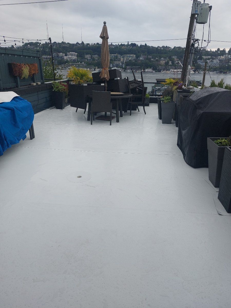 Rooftop patio with gray waterproof coating. Outdoor dining set, umbrella, and planters. Overcast day with city view.