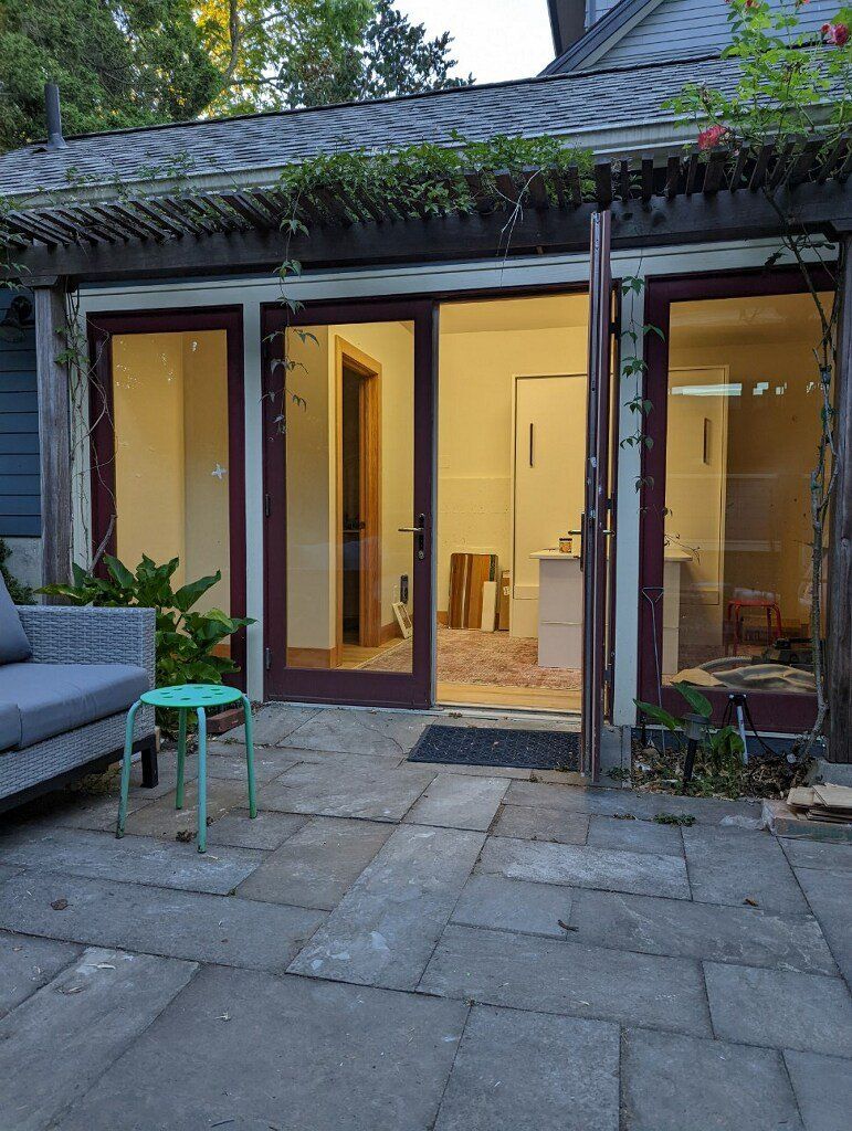 Patio with open glass doors, leading to a warmly lit room. Gray sofa, teal stool, and stone patio.