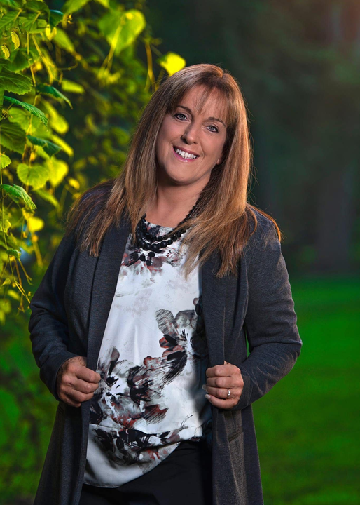 Woman in gray cardigan and floral top smiling, posing outdoors near greenery.