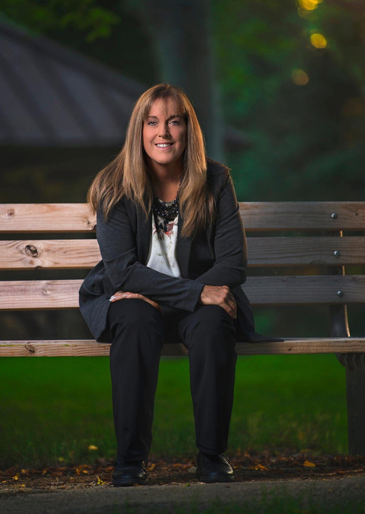 Woman in gray cardigan and floral top smiling, posing outdoors near greenery.