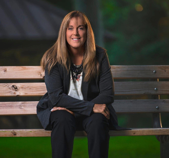 Woman with long brown hair sits on a wooden bench, smiling. She wears a blazer and black pants, outdoors.