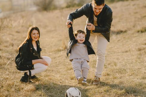 Family playing outdoors: father swings child, mother kneels and smiles. Sunny field.