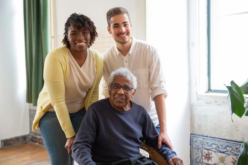 Smiling young man and woman stand behind an elderly man in a wheelchair; all smiling.