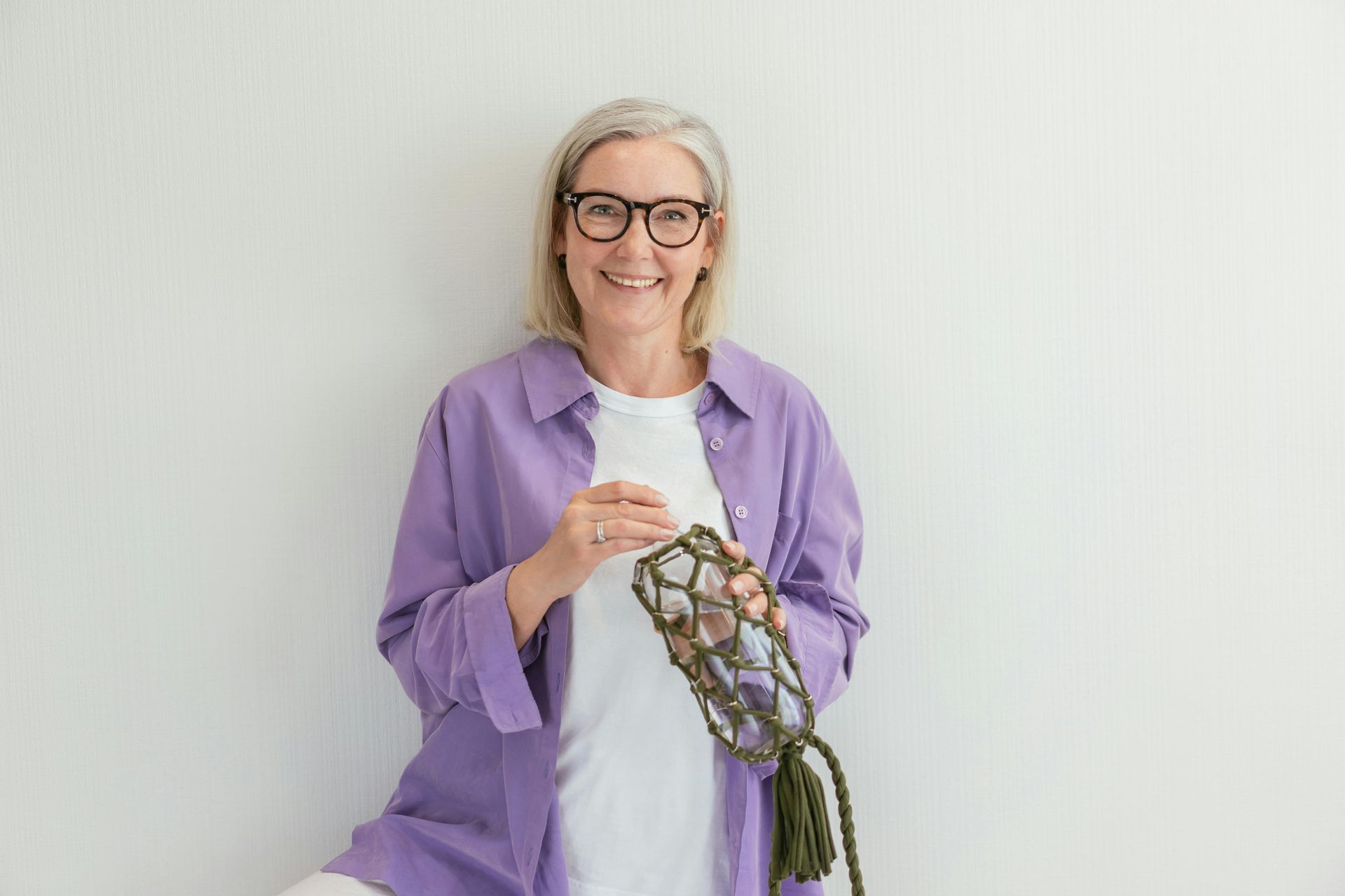 Woman with glasses smiles, crocheting a bottle holder, leaning against a white wall.