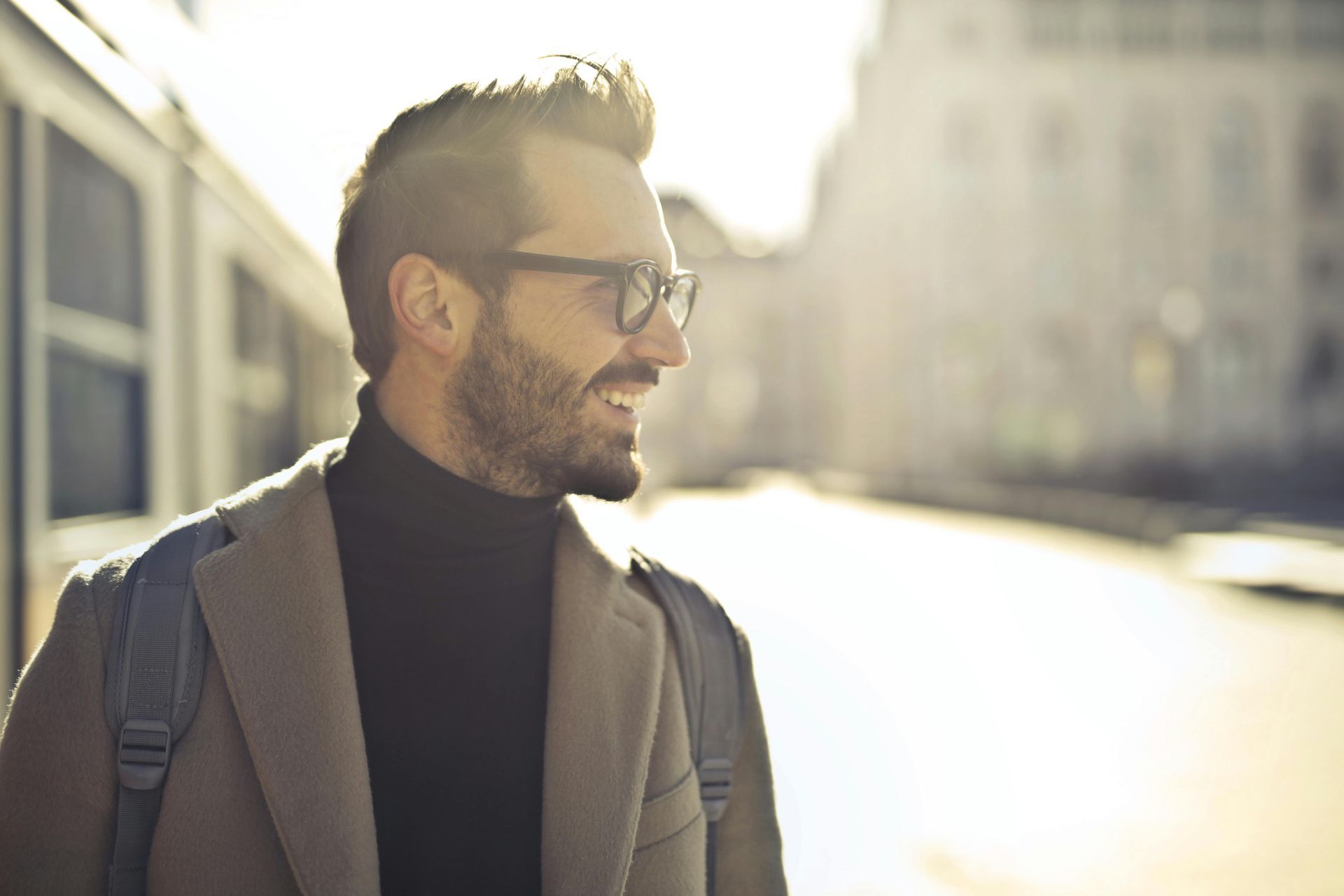 Man with glasses and beard smiles outdoors, wearing a turtleneck and coat, city background.