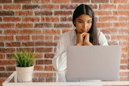 Woman with hands clasped looks at laptop screen, in front of a brick wall.