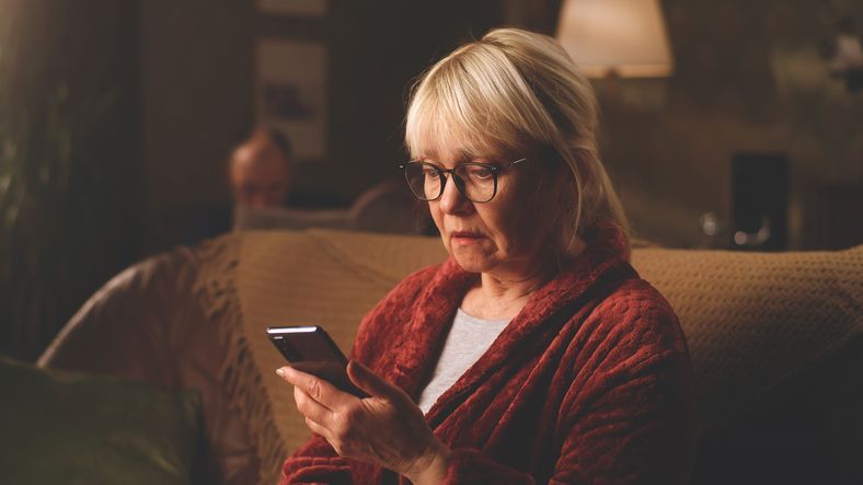 Elderly woman in glasses, red robe, using a smartphone, seated on a couch indoors.