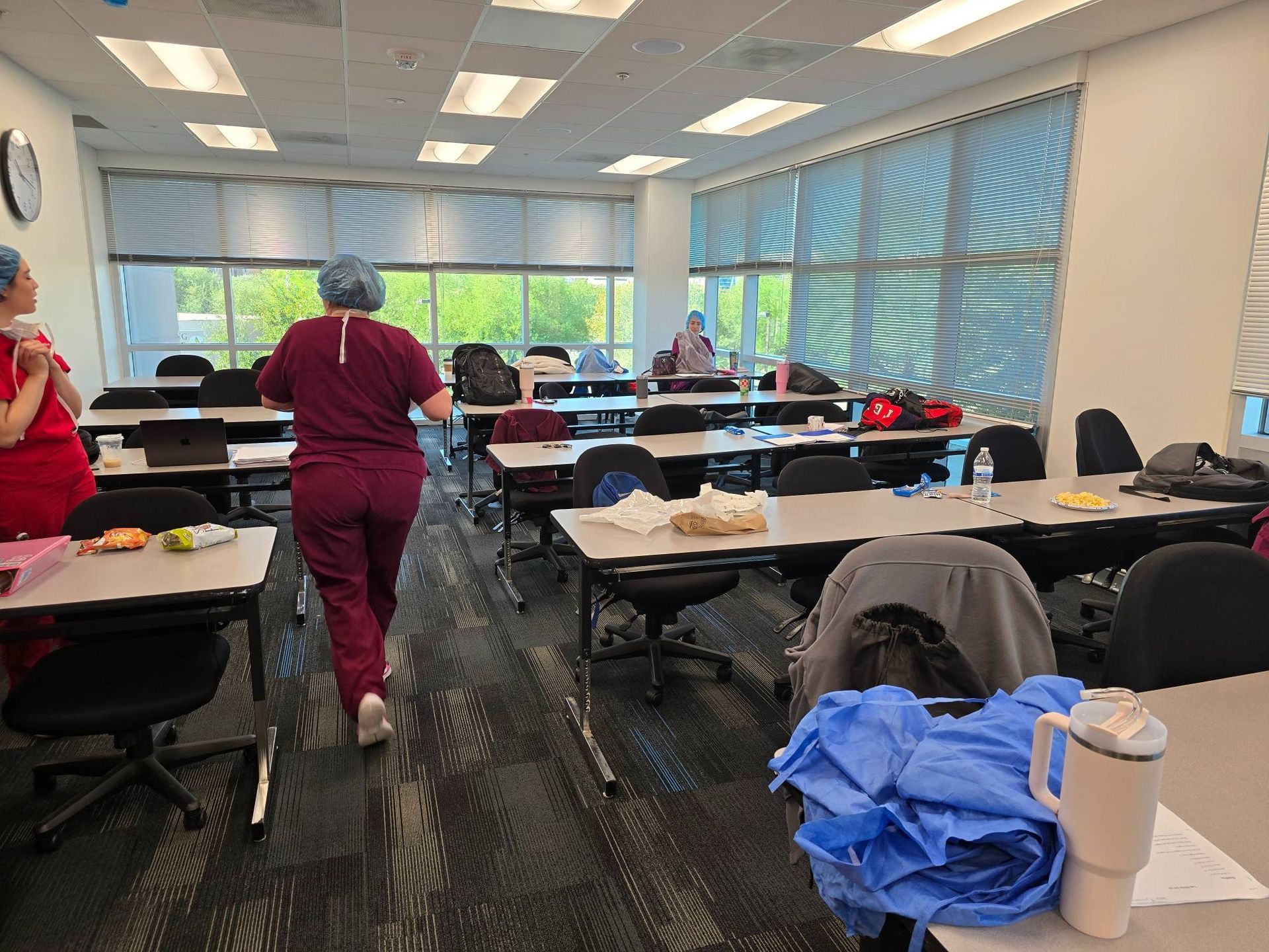 A woman in scrubs is walking through a classroom filled with tables and chairs.