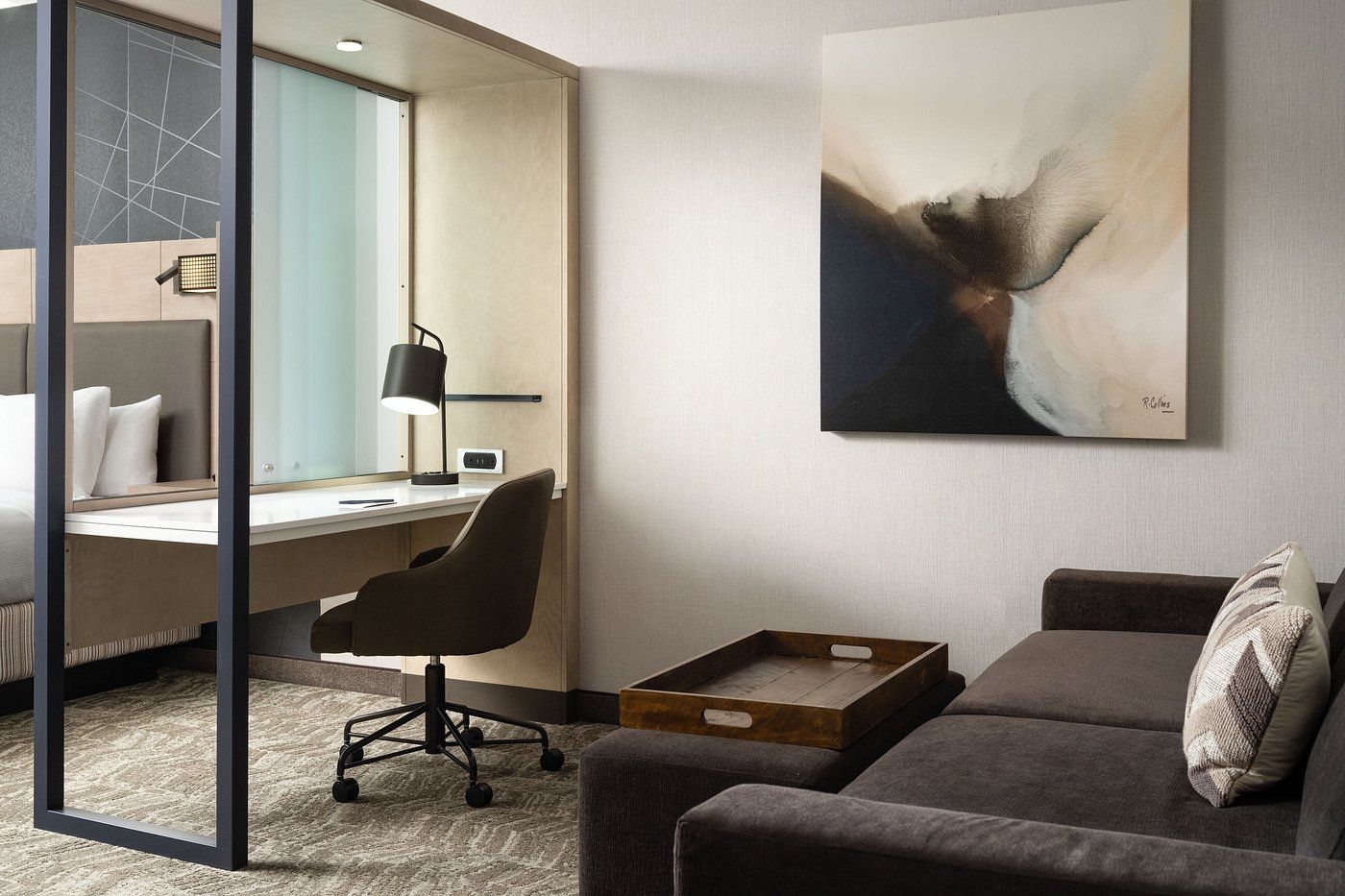 A hotel room with a couch , desk , chair and painting on the wall.