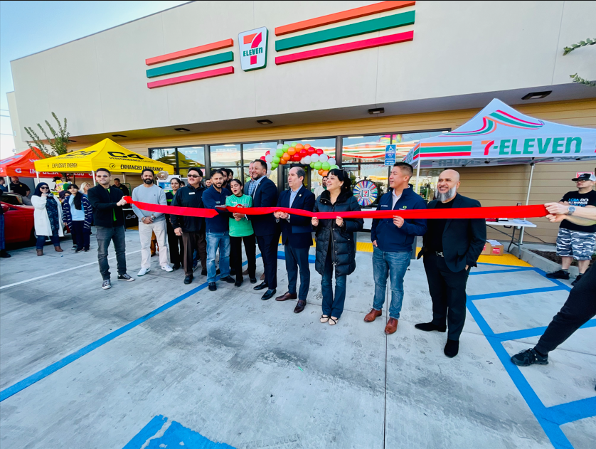 A group of people are cutting a red ribbon in front of a 7 eleven store.