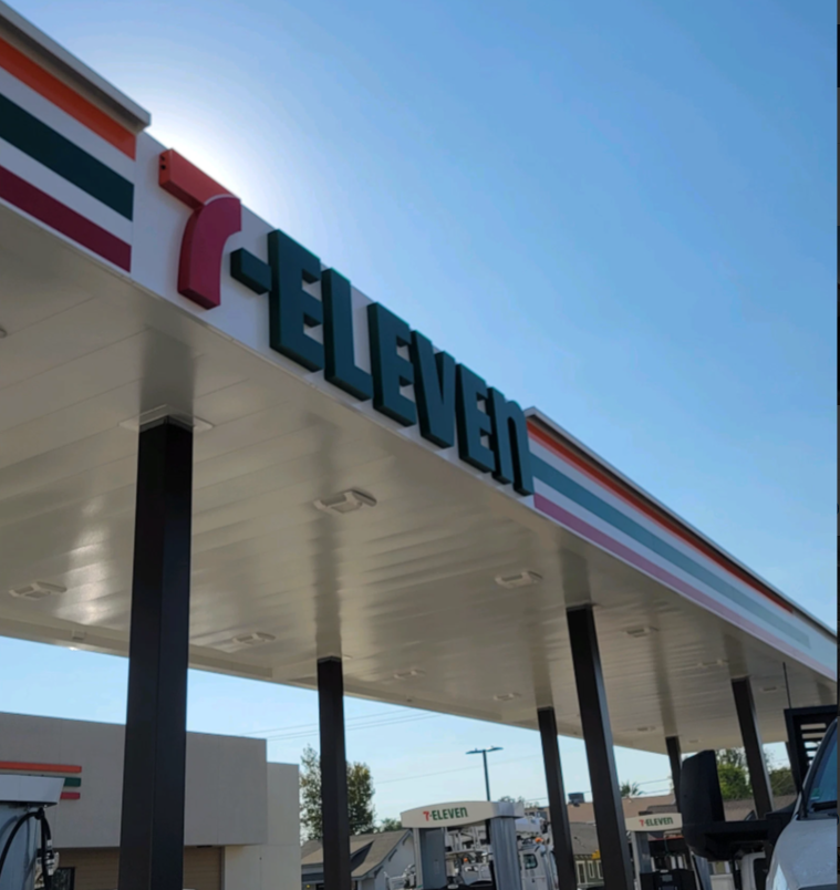 7-Eleven store exterior under a bright blue sky. Overhead awning with sign in red, green, and white.