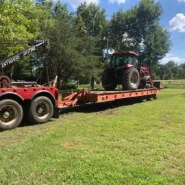 Tractor on a flatbed trailer being towed by a tow truck on grass field.