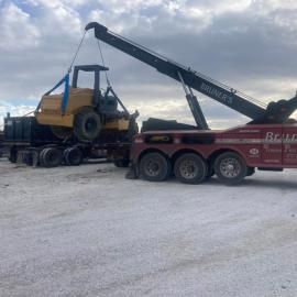 A tow truck lifting a yellow construction machine onto a flatbed trailer. Cloudy sky.
