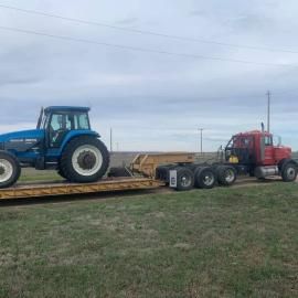 A blue tractor loaded on a flatbed trailer pulled by a red semi-truck on a grassy field.