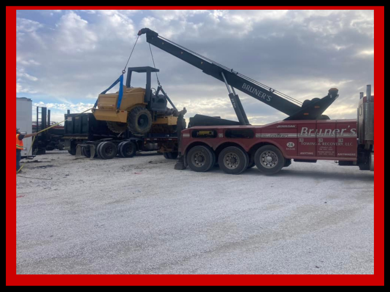 A tow truck lifting a yellow road roller onto a flatbed trailer on a cloudy day.