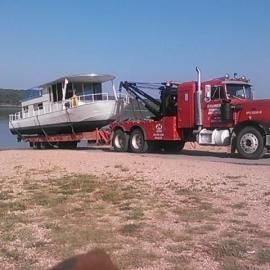 Red tow truck transporting a white houseboat on a trailer near water.