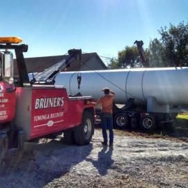 Tow truck lifting a large silver tank on a trailer. A person stands nearby, blue sky overhead.