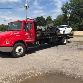 Red tow truck carrying a buggy and a white car on a sunny day.