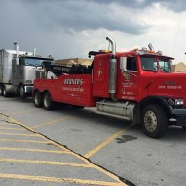 Red tow truck towing a silver semi-truck on a paved lot under a cloudy sky.